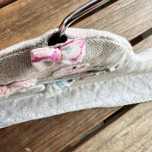 K&ouml;nnte beinhalten: Ein gepolsterter Kleiderb&uuml;gel mit hellbeigem Leinenbezug. Oben ist eine kleine rosa-wei&szlig;e Schleife angebracht. Der B&uuml;gel hat eine Blumenstickerei und eine wei&szlig;e Lochspitze.