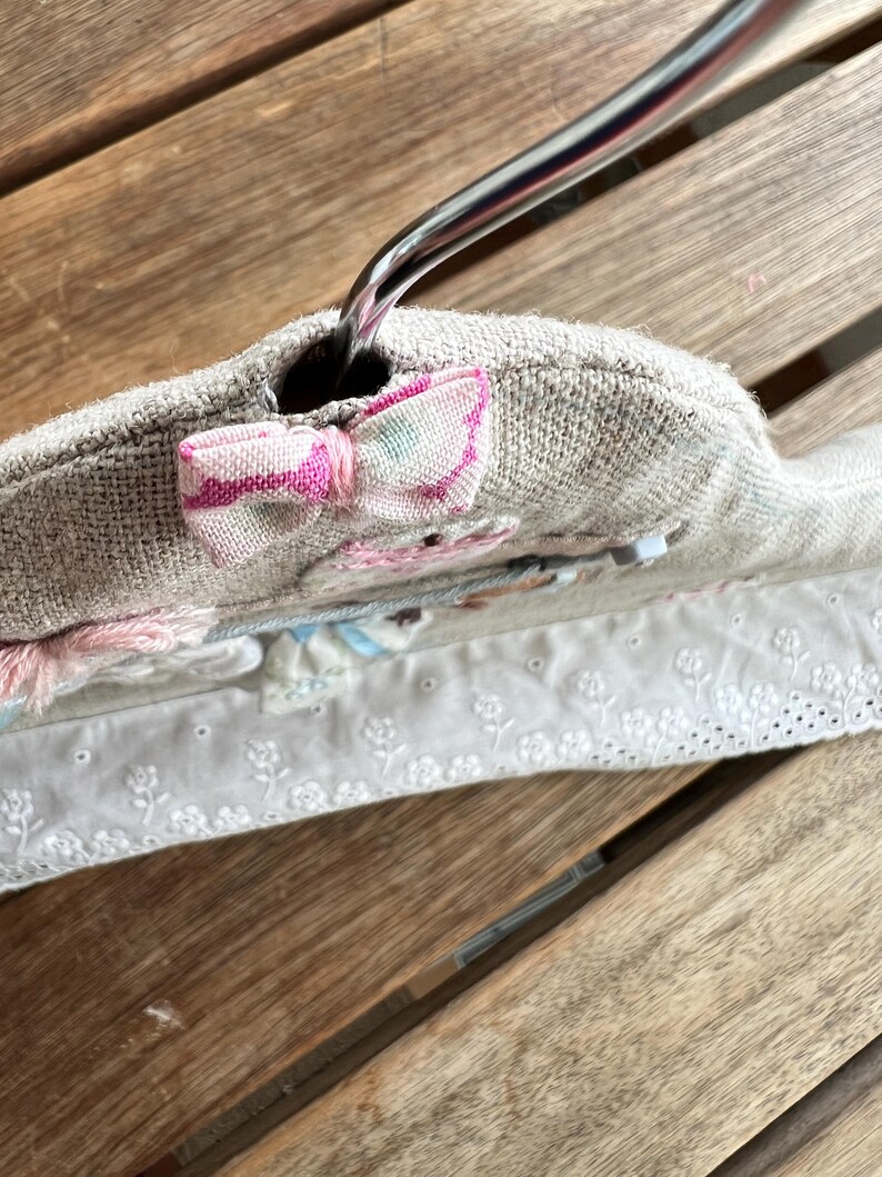 K&ouml;nnte beinhalten: Ein gepolsterter Kleiderb&uuml;gel mit hellbeigem Leinenbezug. Oben ist eine kleine rosa-wei&szlig;e Schleife angebracht. Der B&uuml;gel hat eine Blumenstickerei und eine wei&szlig;e Lochspitze.