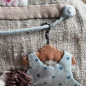 K&ouml;nnte beinhalten: Nahaufnahme eines handgefertigten Textilkunstwerks. Es zeigt ein hellblaues Kleid mit dunkelblauen Punkten, das an einem kleinen braunen Kleiderb&uuml;gel h&auml;ngt. Der Hintergrund ist ein strukturierter, neutralfarbener Stoff mit weiteren Stoffdetails und einem Knopf.