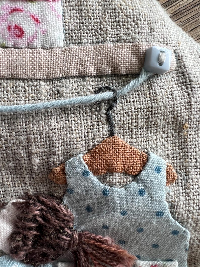 K&ouml;nnte beinhalten: Nahaufnahme eines handgefertigten Textilkunstwerks. Es zeigt ein hellblaues Kleid mit dunkelblauen Punkten, das an einem kleinen braunen Kleiderb&uuml;gel h&auml;ngt. Der Hintergrund ist ein strukturierter, neutralfarbener Stoff mit weiteren Stoffdetails und einem Knopf.