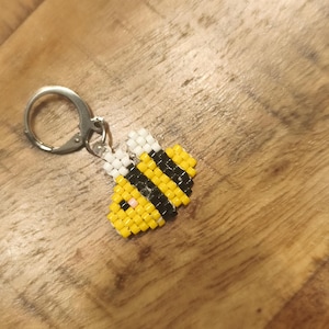 Könnte beinhalten: Ein handgefertigter Bienen-Schlüsselanhänger. Die Biene besteht aus gelben, schwarzen und weißen Perlen mit einem kleinen rosa Detail. Sie ist an einem silberfarbenen Metall-Schlüsselring befestigt. Der Anhänger liegt auf einem Holzgrund.