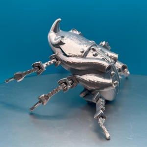 May include: A silver 3D printed model of a beetle with articulated legs. The beetle has a detailed, textured surface and a pointed horn on its head.