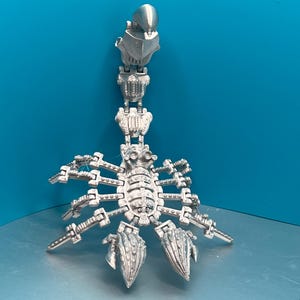 May include: A silver-coloured, articulated scorpion sculpture with a detailed design. The scorpion is posed with its tail raised and its claws extended.
