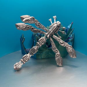 May include: A silver-coloured, 3D printed dragonfly with intricate details and a steampunk aesthetic. The dragonfly is posed with its wings spread out.