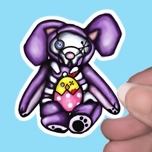 May include: A purple plush bunny rabbit with a skeletal body and a cracked egg with a yellow chick inside. The bunny is wearing a white hood with a button eye and a stitched X over the other eye.