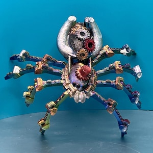 May include: A silver and iridescent steampunk spider sculpture with gears and cogs. The spider has eight legs and a round body with a silver and iridescent finish.
