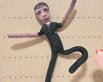 May include: A fabric doll featuring a printed human face with short dark hair, wearing a black scrub top with a printed stethoscope. The doll has long, thin arms and legs with visible stitching along the edges, posed in a dynamic, outstretched position.