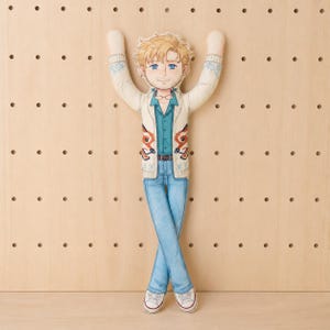 May include: A flat fabric doll featuring a cartoon illustration of a person with blonde hair and blue eyes. The figure wears a cream-colored cardigan with fox motifs, a teal button-down shirt, blue jeans, and white sneakers with arms raised.