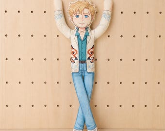 May include: A flat fabric doll featuring a cartoon illustration of a person with blonde hair and blue eyes. The figure wears a cream-colored cardigan with fox motifs, a teal button-down shirt, blue jeans, and white sneakers with arms raised.
