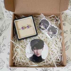 May include: A cardboard gift box filled with shredded paper containing a pair of heart-shaped stud earrings in a black display box, two small anime-style character stickers, and a circular pinback button featuring a portrait of a person with dark hair and the text Calab.