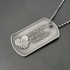 May include: A brushed silver-tone metal dog tag pendant on a matching ball chain. The tag features an embossed graphic of two hands forming a heart shape and the engraved text LIGHTFOOT MEDICAL TRANSPORT in a clean, sans-serif capital font.