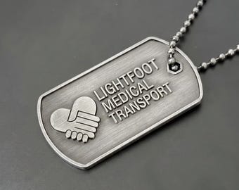 May include: A brushed silver-tone metal dog tag pendant on a matching ball chain. The tag features an embossed graphic of two hands forming a heart shape and the engraved text LIGHTFOOT MEDICAL TRANSPORT in a clean, sans-serif capital font.