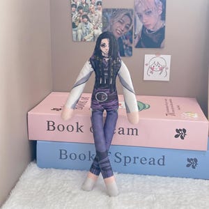 以下が含まれることがあります： 長い黒髪のキャラクターがプリントされた布製のぬいぐるみ。ベルト付きの紫と白の衣装を着ている。細身の体型で、長い腕を持ち、足首を交差させて立っている。キャラクターは大きな目と黒髪が特徴。