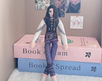 May include: A plush doll featuring a printed character with long black hair, wearing a purple and white outfit with a belt. The doll has a slender build, long arms, and legs crossed at the ankles. The character has large eyes and dark hair.