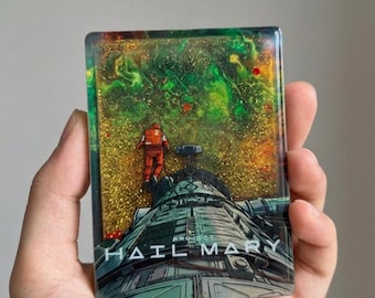 May include: A rectangular resin art piece featuring a space scene with a metallic gray spaceship and an astronaut in an orange suit floating against a background of swirling green and gold glitter. The text PROJECT HAIL MARY is printed in white.