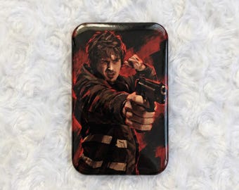 May include: A rectangular pinback button featuring a stylized illustration of a person with dark hair wearing a dark jacket, holding a black handgun pointed forward. The background consists of bold, expressive red and black brushstroke textures.