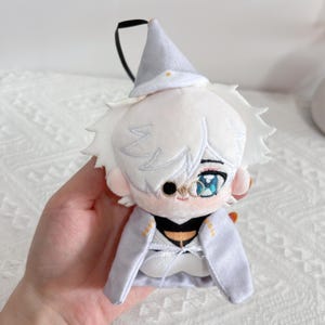 May include: A plush doll featuring white hair, a pointed hat, and a monocle over one eye. The doll wears a light gray cape with gold accents and a white ribbon tie. It has large, detailed blue eyes and a small embroidered mouth, with a black loop attached.