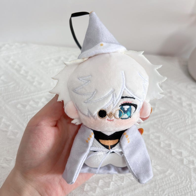 May include: A plush doll featuring white hair, a pointed hat, and a monocle over one eye. The doll wears a light gray cape with gold accents and a white ribbon tie. It has large, detailed blue eyes and a small embroidered mouth, with a black loop attached.