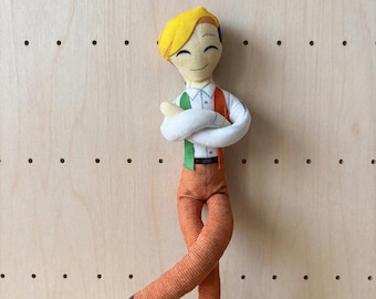 May include: A fabric doll with yellow hair, a white collared shirt, green and orange suspenders, and brown trousers. The doll has a smiling expression with closed eyes, arms crossed over the chest, and legs positioned in a crossed stance.
