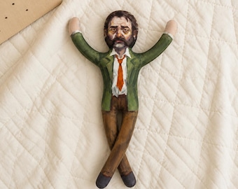 May include: A fabric doll featuring a printed illustration of a person with a mustache and dark hair, wearing a green blazer, white shirt, orange tie, and brown trousers. The doll has arms raised upward and legs crossed at the ankles.