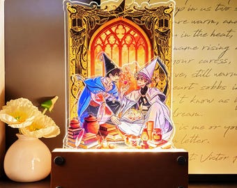 May include: A rectangular acrylic light display featuring an illustration of two figures in pointed hats and robes sitting amidst books and jars. The scene is framed by an ornate golden archway. The display sits on a dark wooden base with four feet.