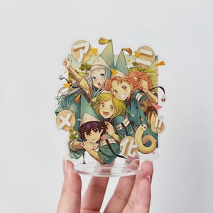 May include: A clear acrylic standee featuring an anime-style illustration of four characters wearing pointed hats and teal outfits. The design includes Japanese text in circular speech bubbles, colorful confetti accents, and a landscape background.