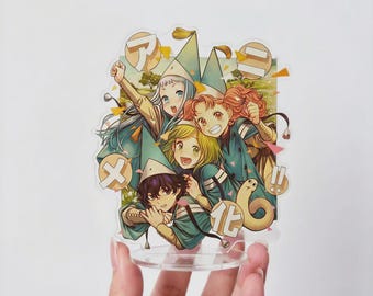 May include: A clear acrylic standee featuring an anime-style illustration of four characters wearing pointed hats and teal outfits. The design includes Japanese text in circular speech bubbles, colorful confetti accents, and a landscape background.