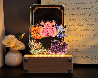 May include: A rectangular acrylic light display featuring five colorful anime-style character illustrations layered on a dark background. The clear acrylic panel is mounted on a rectangular wooden base with four silver circular accents at the corners.