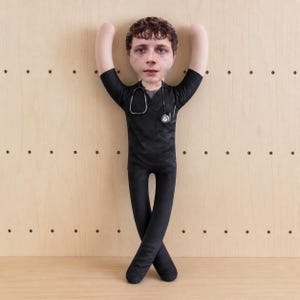 Puede incluir: Una muñeca de tela personalizada con la fotografía impresa de un rostro con cabello castaño. La muñeca viste una casaca de cuello en V negra con un estetoscopio impreso alrededor del cuello y pantalones negros a juego, con los brazos levantados.