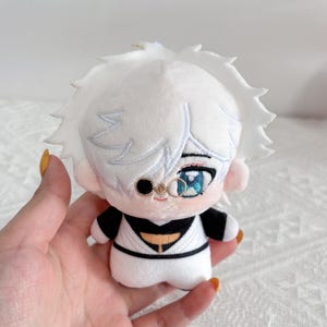 May include: A plush doll featuring a character with short white hair, one black circular monocle over the right eye, and a blue iris in the left eye. The doll wears a white outfit with black accents, including a black collar and a black belt detail.