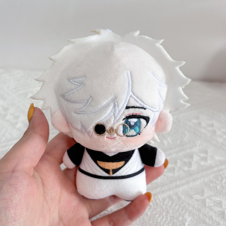 May include: A plush doll featuring a character with short white hair, one black circular monocle over the right eye, and a blue iris in the left eye. The doll wears a white outfit with black accents, including a black collar and a black belt detail.