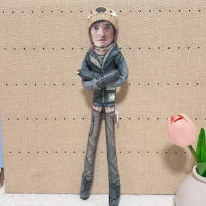 May include: A flat, fabric-printed doll featuring a person with a realistic face wearing a tan beanie with sheep graphics, a dark blue jacket with a white cross emblem, and gray pants. The doll has arms crossed and a zipper visible on the side.