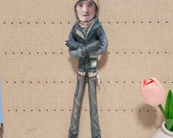 May include: A flat, fabric-printed doll featuring a person with a realistic face wearing a tan beanie with sheep graphics, a dark blue jacket with a white cross emblem, and gray pants. The doll has arms crossed and a zipper visible on the side.
