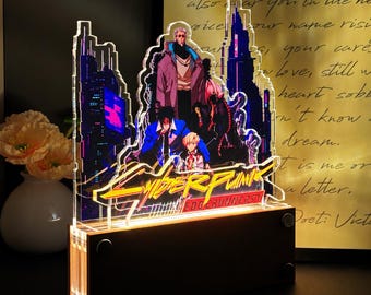 May include: A multi-layered acrylic light display featuring Cyberpunk Edgerunners characters and city silhouettes. The design includes a yellow and red logo, illuminated by a warm base. The clear acrylic panels show detailed anime-style figures against a dark, neon-lit urban background, mounted on a rectangular wooden stand.