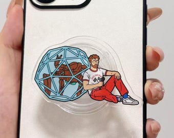 May include: A clear circular phone grip featuring a graphic of a person with a beard and glasses wearing a white t-shirt with the text Horse Shoe Bend and red pants, sitting next to a large blue geometric sphere containing a brown crab-like creature.