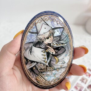 May include: An oval-shaped decorative pin featuring an anime-style illustration of a character with light hair wearing a white hat and cape with gold buttons. The design includes intricate gold filigree borders and the text "Ollrp" in gold script.