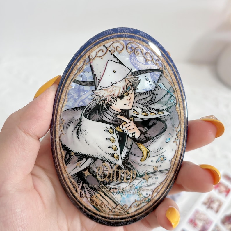 May include: An oval-shaped decorative pin featuring an anime-style illustration of a character with light hair wearing a white hat and cape with gold buttons. The design includes intricate gold filigree borders and the text "Ollrp" in gold script.