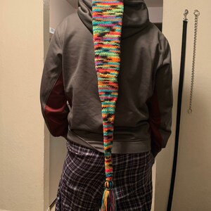 May include: A long, colorful knit hat with a pointed end and a tassel. The hat features a vibrant mix of colors including blue, green, orange, and pink. The hat is worn by a person wearing a gray hoodie and plaid pants.