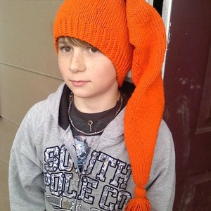 May include: An orange knit hat with a long, pointed end and a tassel. The hat is a vibrant orange color and appears to be handmade. The hat is worn by a person wearing a gray hoodie.