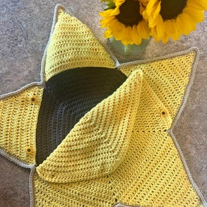 May include: A handmade, star-shaped crochet item in shades of yellow and gray. The item has a sunflower design with a gray border and small sunflower accents. The center is a darker gray, and the edges are a lighter yellow.