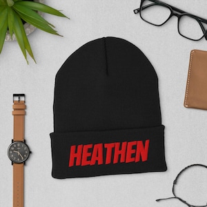 以下が含まれることがあります： 赤い文字で「HEATEN」と刺繍された黒いビーニー帽。帽子はつばが折り返されています。画像には、時計、メガネ、小さな財布などのアイテムも含まれています。