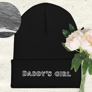 May include: Black beanie with the text "DADDY'S GIRL" embroidered in white. The hat is made of a soft, knitted material. A bouquet of white flowers with green leaves is in the background.