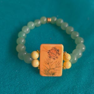May include: A stretch bracelet featuring a series of smooth, round, light green beads. The center focal point is a rectangular, cream-colored tile with a black line drawing of a flower and a small insect. Four yellow beads flank the central tile.