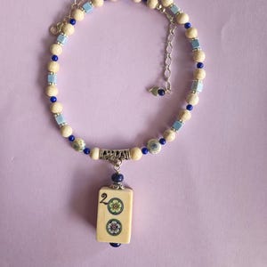 May include: A necklace featuring a central rectangular mahjong tile pendant with a number two and two floral designs. The chain consists of round cream-colored beads, small blue cube beads, tiny dark blue spheres, and a silver-toned metal extender chain.