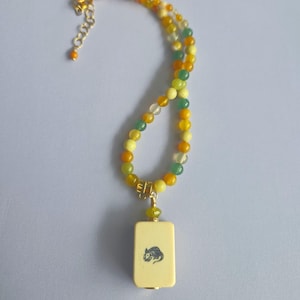 May include: A necklace featuring a strand of round beads in shades of yellow, orange, and green, accented with gold-toned metal hardware. A rectangular, cream-colored pendant hangs from the center, displaying a small, dark illustration of a rat.