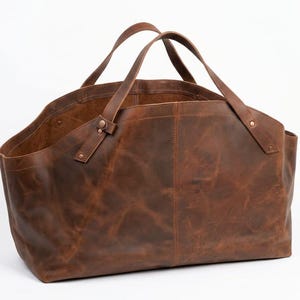 Könnte beinhalten: Große, offene braune Leder-Tragetasche mit zwei stabilen Griffen. Die Tasche hat eine Used-Optik, sichtbare Nähte und ein schlichtes Design. Die Griffe sind mit Nieten befestigt.