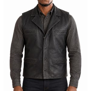 May include: A black leather vest featuring a notched lapel collar, a front button closure, and four horizontal slit pockets. The vest is worn over a dark gray long-sleeved button-down shirt and dark denim jeans, creating a layered look.