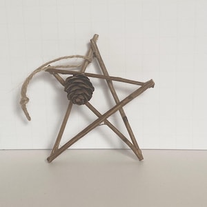 May include: A rustic star-shaped ornament crafted from twigs, featuring a small pinecone at its center. A natural twine loop is attached for hanging. The brown tones of the wood and pinecone create a warm, earthy aesthetic, perfect for holiday decor.