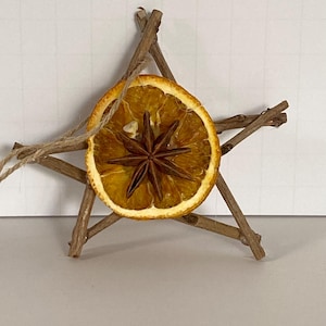 Może przedstawiać: Ozdoba w kształcie gwiazdy wykonana z gałązek, plasterka suszonej pomarańczy i anyżu gwiaździstego. Do zawieszenia przymocowana jest pętla ze sznurka. Plaster pomarańczy jest wyśrodkowany, a anyż gwiaździsty w środku.
