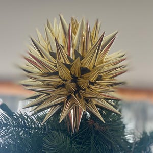 May include: A gold starburst Christmas tree topper with sharp, spiky points. The starburst is metallic gold with red accents and sits atop a green Christmas tree. The starburst is a decorative holiday ornament.